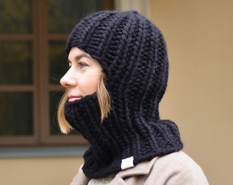 May include: A black knitted balaclava with a long neck covering the face and neck. The balaclava is worn by a person with a brown coat.