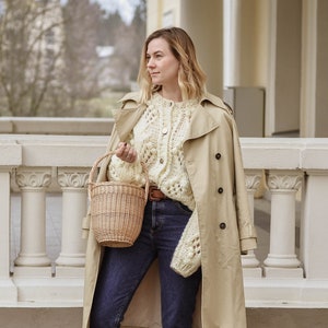 May include: A woman wearing a beige trench coat, cream-colored cable-knit sweater, and dark blue jeans. She holds a small woven basket and wears black loafers. The background features a stone walkway and a white building.