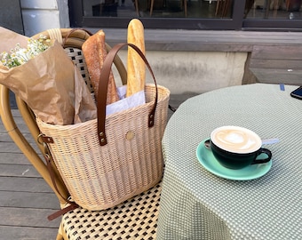 May include: A wicker basket with a leather handle contains two baguettes and a bouquet of flowers. A cup of coffee sits on a green saucer on a table with a green and white patterned tablecloth. The scene is outdoors.