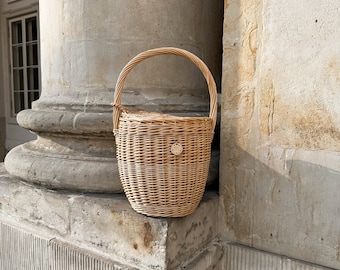 May include: A light brown wicker basket with a rounded body and a curved handle. The basket has a small, circular emblem on the front. The basket is sitting on a stone ledge next to a large column.