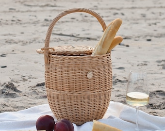 May include: A wicker picnic basket with a lid and a handle, filled with baguettes, on a white blanket on a sandy beach. There are two red plums, two containers of cream cheese, and a glass of white wine on the blanket.