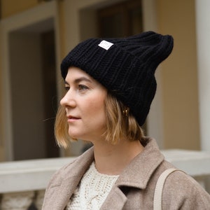 May include: A woman wearing a black knit beanie with a white logo. She is wearing a beige coat and has short blonde hair.