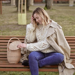 May include: A light beige, round wicker basket with a lid and handle sits on a wooden bench. The basket is next to a person wearing a cream-colored, open-knit sweater, blue jeans, and a tan trench coat. The background is a park setting.