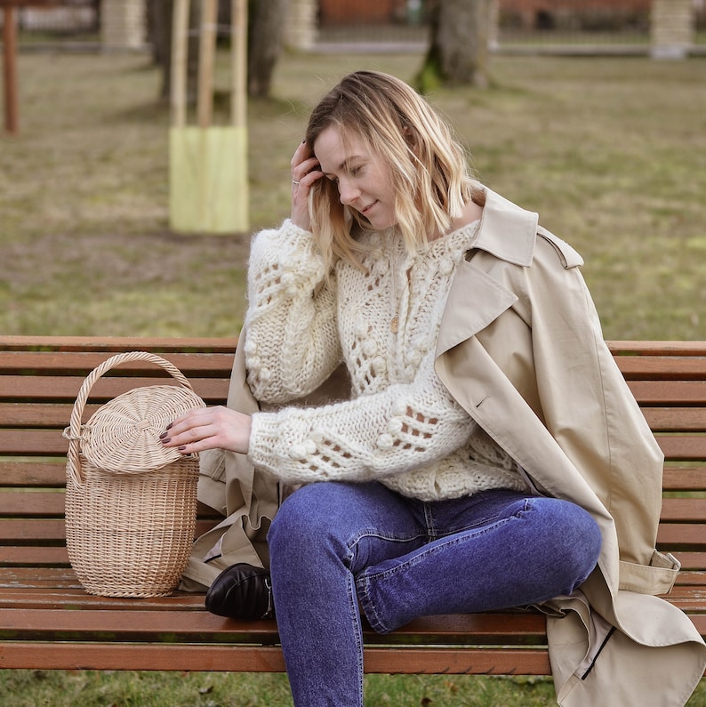 May include: A light beige, round wicker basket with a lid and handle sits on a wooden bench. The basket is next to a person wearing a cream-colored, open-knit sweater, blue jeans, and a tan trench coat. The background is a park setting.