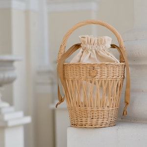 May include: A woven wicker basket with a rounded handle and tan fabric lining. The basket has a drawstring closure and tan straps. A small wooden emblem with the letter "m" is attached to the front.