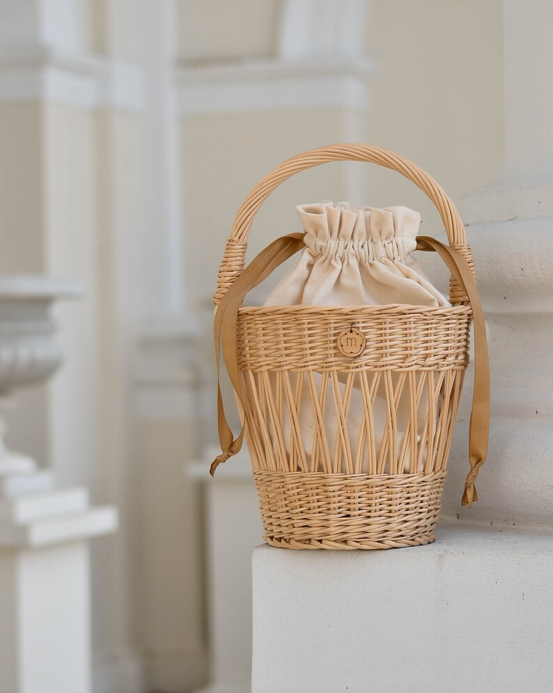 May include: A woven wicker basket with a rounded handle and tan fabric lining. The basket has a drawstring closure and tan straps. A small wooden emblem with the letter "m" is attached to the front.