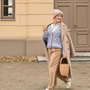 May include: A woman wearing a light blue cable knit cardigan with a matching beanie, tan pants, and a tan coat. She carries a small woven basket. The background features a building with a tan facade and a brown door.