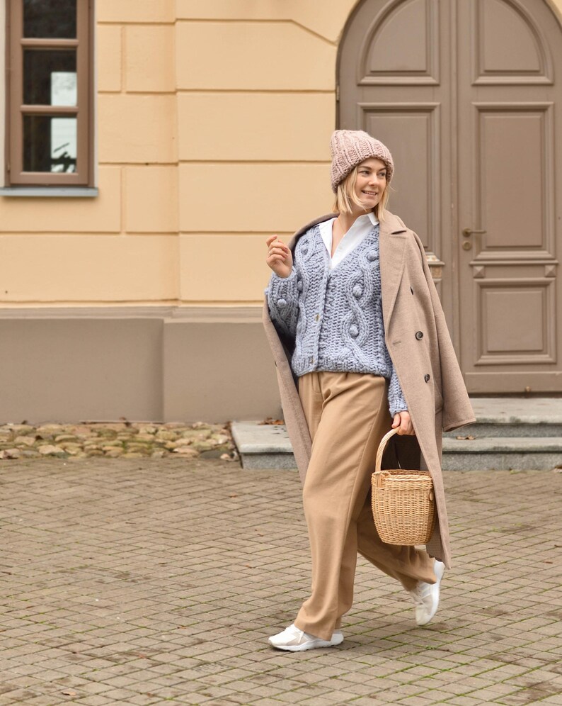 May include: A woman wearing a light blue cable knit cardigan with a matching beanie, tan pants, and a tan coat. She carries a small woven basket. The background features a building with a tan facade and a brown door.