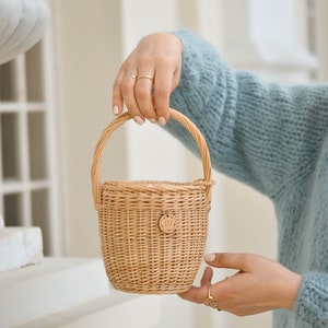 May include: A small, woven wicker basket with a rounded handle. The basket is light brown with a circular wooden tag that has the letter "m" on it. The basket is held by a person wearing a light blue jumper.