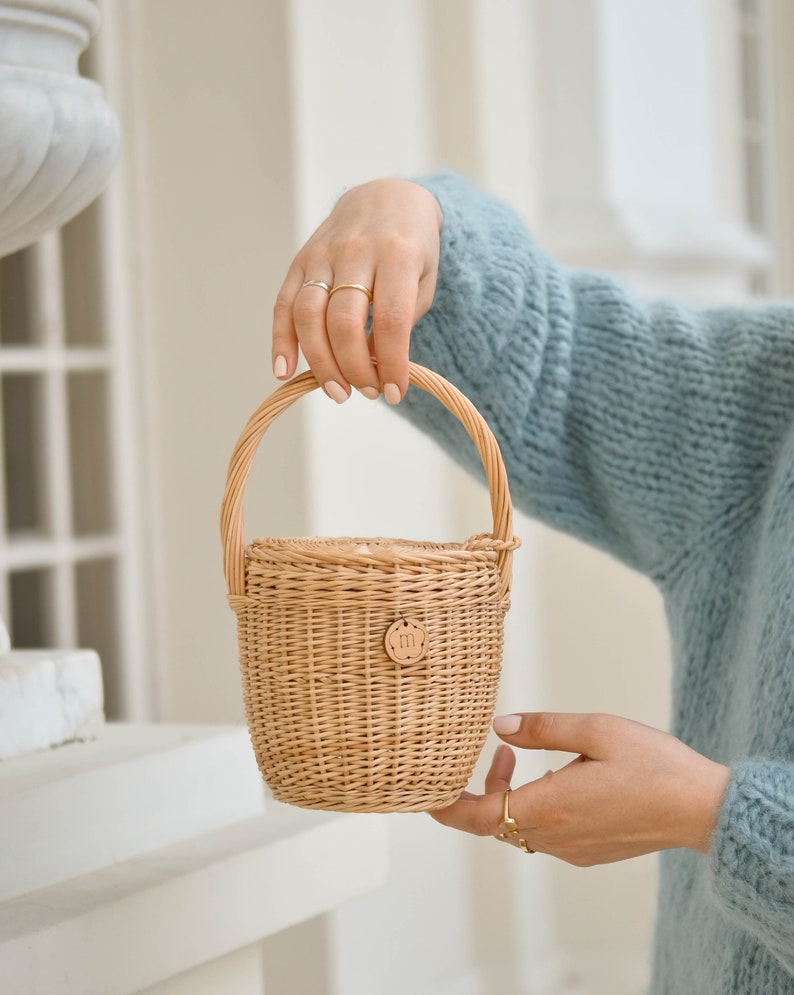 May include: A small, woven wicker basket with a rounded handle. The basket is light brown with a circular wooden tag that has the letter "m" on it. The basket is held by a person wearing a light blue jumper.