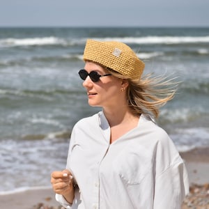 May include: A mustard yellow crocheted hat with a square crown and a small white label. The hat is worn with black sunglasses and a white button-down shirt. The background shows the ocean and a sandy beach.