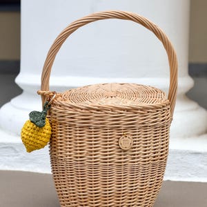 May include: A woven wicker basket with a rounded shape, a handle, and a lid. A small, yellow crocheted lemon charm with green leaves hangs from the handle. A small wooden tag with the letter "m" is attached to the basket.