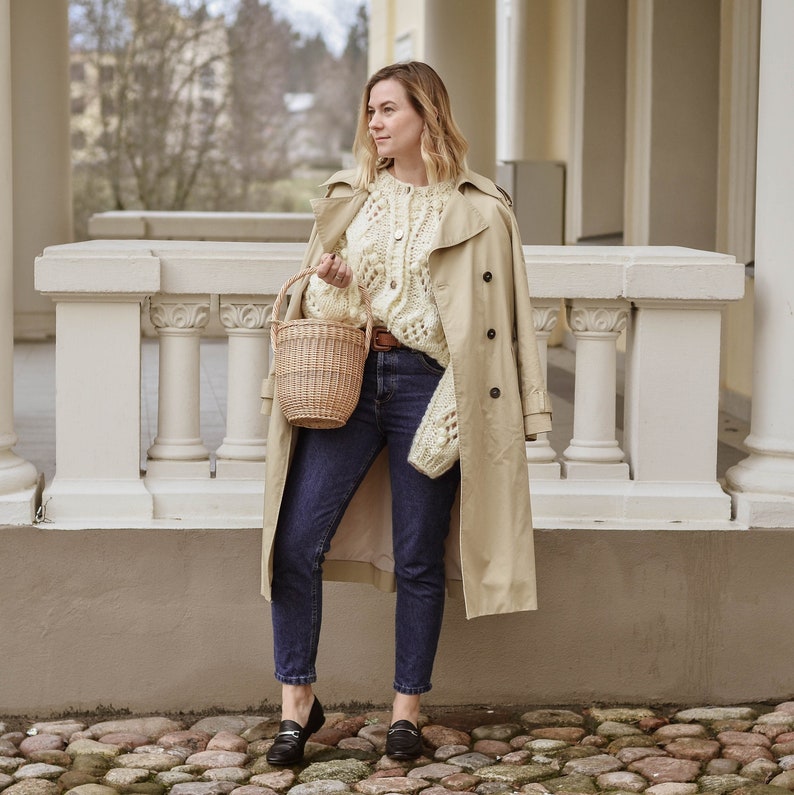 May include: A woman wearing a beige trench coat, cream-colored cable-knit sweater, and dark blue jeans. She holds a small woven basket and wears black loafers. The background features a stone walkway and a white building.