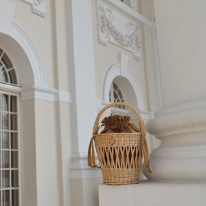 May include: A woven wicker basket with a brown drawstring pouch inside. The basket has a handle and brown straps. The basket is sitting on a white surface in front of a building with white columns and windows.
