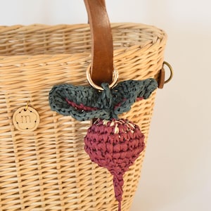 May include: A woven wicker basket with a brown leather handle and gold-colored hardware. A decorative charm in the shape of a beet is attached to the basket. The beet charm is a deep red color with a green leaf accent.