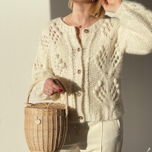 May include: A cream-colored, open-knit cardigan with button closures and a round neckline. The cardigan is paired with cream-colored pants. A small, woven basket with a handle is held in the hand.