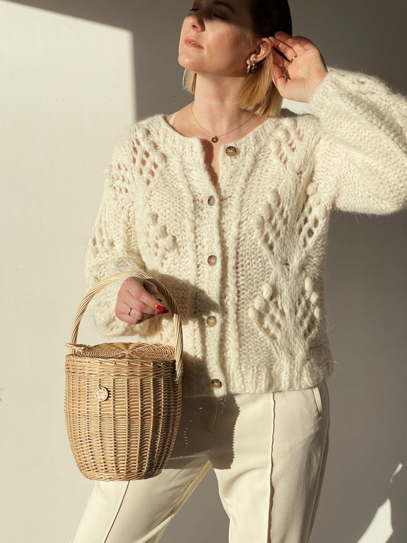 May include: A cream-colored, open-knit cardigan with button closures and a round neckline. The cardigan is paired with cream-colored pants. A small, woven basket with a handle is held in the hand.