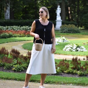 May include: A woman wearing a black crocheted sleeveless top with a white skirt and white shoes. She is carrying a small woven basket bag.