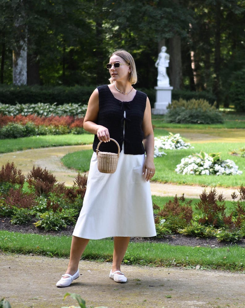 May include: A woman wearing a black crocheted sleeveless top with a white skirt and white shoes. She is carrying a small woven basket bag.