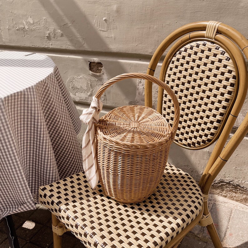 May include: A light brown wicker picnic basket with a handle and a lid, sitting on a chair with a woven seat. A striped cloth is tied to the handle. A table with a checkered tablecloth is in the background.