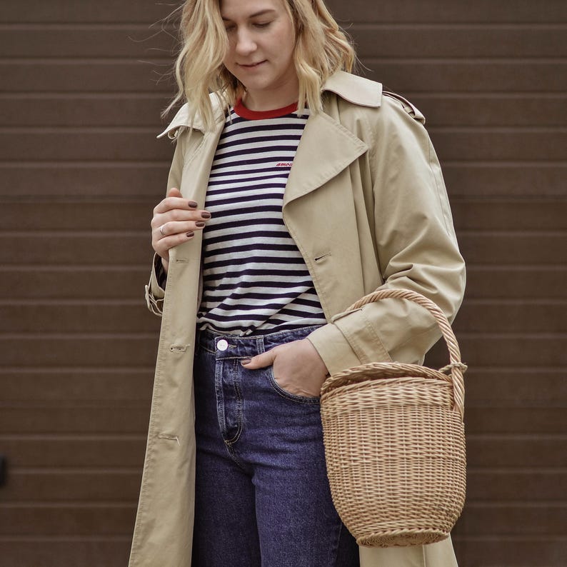 May include: A woman wearing a tan trench coat, blue jeans, and a striped shirt with a red collar. She is holding a woven basket bag with a handle. The background is a brown garage door.