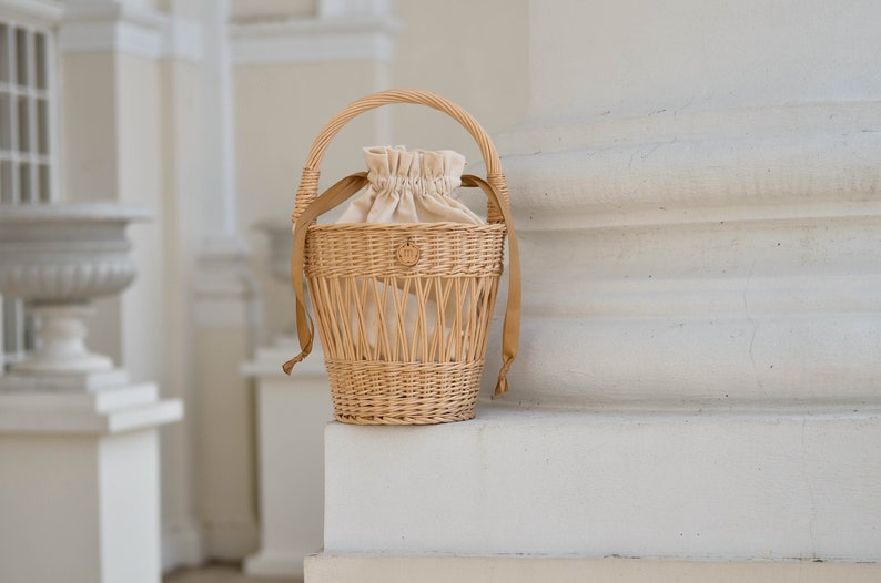 May include: A light brown wicker basket with a handle and a beige drawstring bag inside. The basket has a woven design and tan ribbons. The basket is sitting on a white surface.