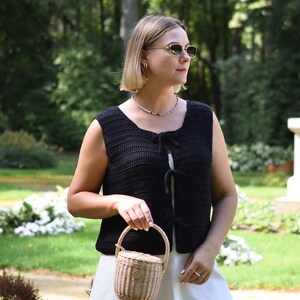 May include: A woman wearing a black crochet vest with a white dress and a small wicker basket bag. She is wearing sunglasses and a beaded necklace.