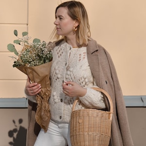 May include: A woman wearing a cream-colored, open-knit cardigan with a brown coat draped over her shoulders. She holds a bouquet of flowers wrapped in brown paper and a woven basket. She is wearing gold hoop earrings and white jeans.