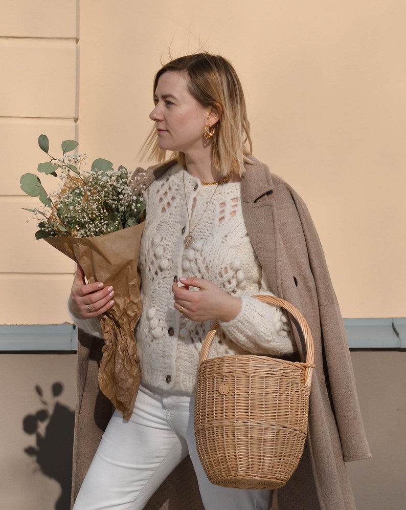 May include: A woman wearing a cream-colored, open-knit cardigan with a brown coat draped over her shoulders. She holds a bouquet of flowers wrapped in brown paper and a woven basket. She is wearing gold hoop earrings and white jeans.