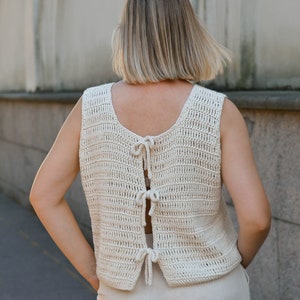 May include: A crocheted sleeveless top with a tie back detail. The top is made of a cream-colored yarn and has a loose, relaxed fit.