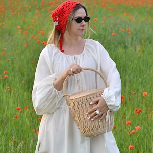 May include: A woman in a field of red poppies wears a red crocheted head covering and black sunglasses. She holds a woven basket and is dressed in a white dress with long sleeves. The scene is set in a field of green grass and red flowers.
