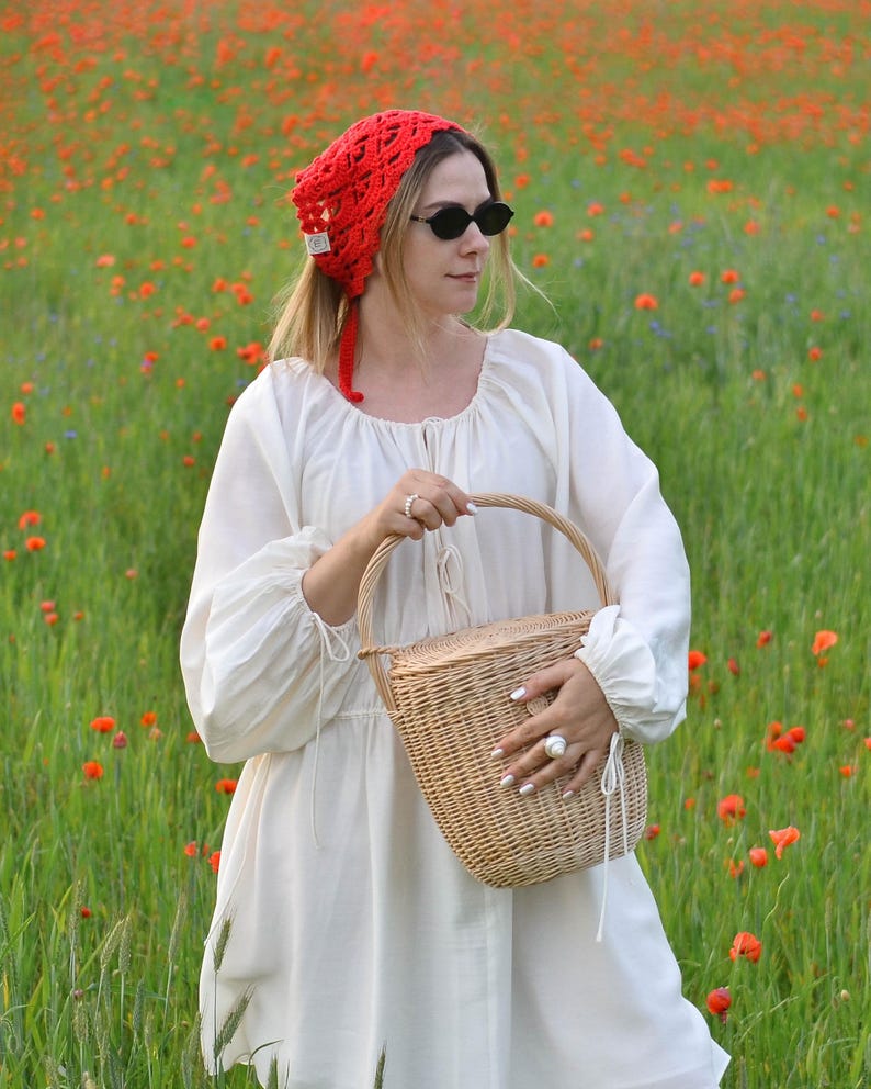 May include: A woman in a field of red poppies wears a red crocheted head covering and black sunglasses. She holds a woven basket and is dressed in a white dress with long sleeves. The scene is set in a field of green grass and red flowers.