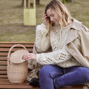 May include: A light brown woven wicker basket with a handle and a lid. The basket is round and has a textured surface. The person is wearing a cream-coloured sweater, blue jeans, and a tan trench coat.