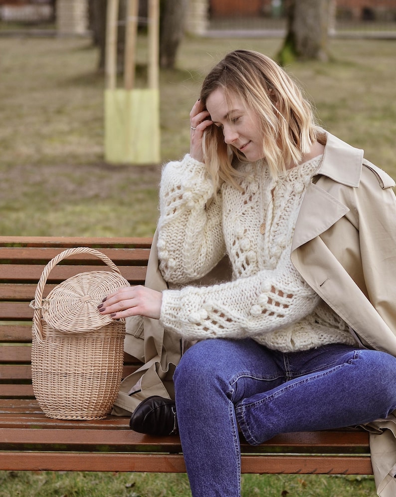 May include: A light brown woven wicker basket with a handle and a lid. The basket is round and has a textured surface. The person is wearing a cream-coloured sweater, blue jeans, and a tan trench coat.