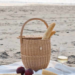 May include: A wicker picnic basket with a handle, filled with baguettes, sits on a white blanket on a sandy beach. Two ripe peaches, cheese, and a glass of white wine complete the scene. The basket has a round lid and a small decorative emblem.