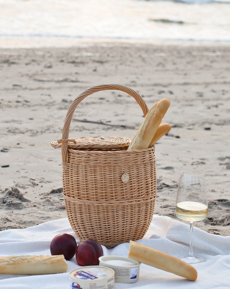 May include: A wicker picnic basket with a handle, filled with baguettes, sits on a white blanket on a sandy beach. Two ripe peaches, cheese, and a glass of white wine complete the scene. The basket has a round lid and a small decorative emblem.