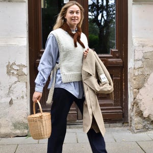 May include: A woman wearing a cream-colored knit vest over a blue striped shirt and dark blue pants. She carries a woven basket and a tan trench coat. A brown scarf is around her neck. The background features a wooden door.