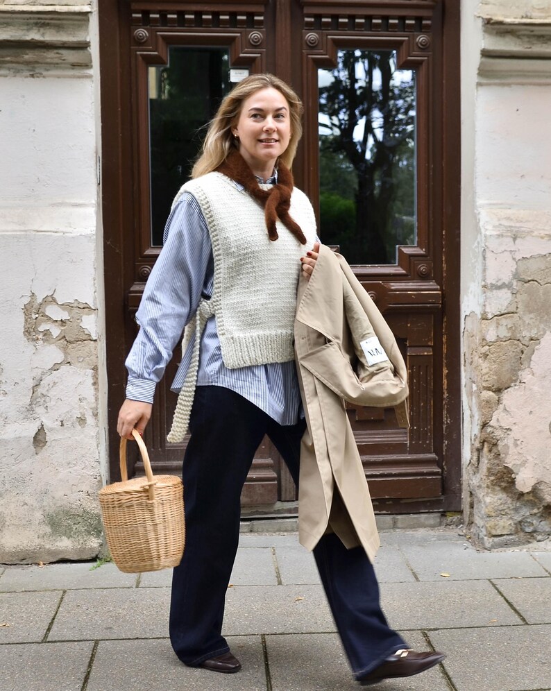 May include: A woman wearing a cream-colored knit vest over a blue striped shirt and dark blue pants. She carries a woven basket and a tan trench coat. A brown scarf is around her neck. The background features a wooden door.