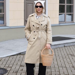 May include: A tan trench coat with a matching belt, black pants, and a white cat-ear hat. The person is holding a woven basket. The outfit is completed with black sunglasses and sneakers.