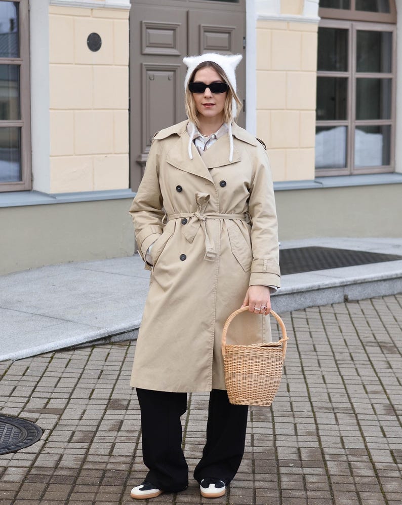 May include: A tan trench coat with a matching belt, black pants, and a white cat-ear hat. The person is holding a woven basket. The outfit is completed with black sunglasses and sneakers.