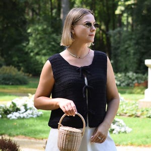 Pu&ograve; includere: Un gilet nero all'uncinetto con chiusure a fiocco &egrave; indossato sopra un vestito bianco. Si tiene un piccolo cestino intrecciato con manico. Lo sfondo presenta un prato verde e alberi.
