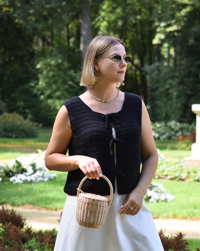 Pu&ograve; includere: Un gilet nero all'uncinetto con chiusure a fiocco &egrave; indossato sopra un vestito bianco. Si tiene un piccolo cestino intrecciato con manico. Lo sfondo presenta un prato verde e alberi.