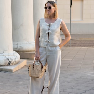 May include: A woman wearing a white crocheted vest with a tie detail, white wide-leg pants, and sunglasses. She is carrying a woven straw bag with a leather strap.