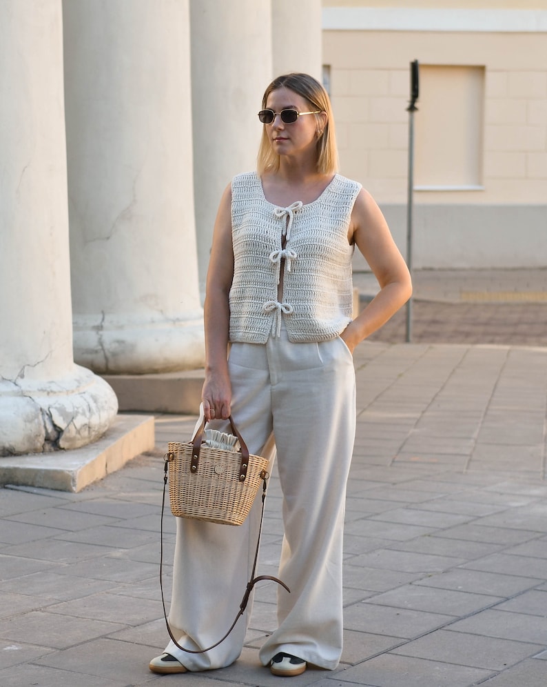 May include: A woman wearing a white crocheted vest with a tie detail, white wide-leg pants, and sunglasses. She is carrying a woven straw bag with a leather strap.