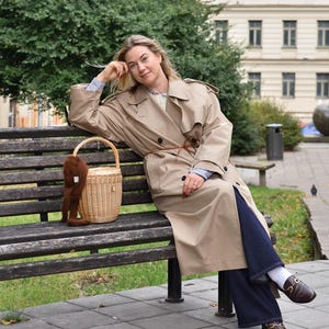 May include: A woman wearing a tan trench coat, blue jeans, and brown loafers sits on a wooden bench. A woven basket with a brown furry accessory rests beside her. The background includes a building and green trees.