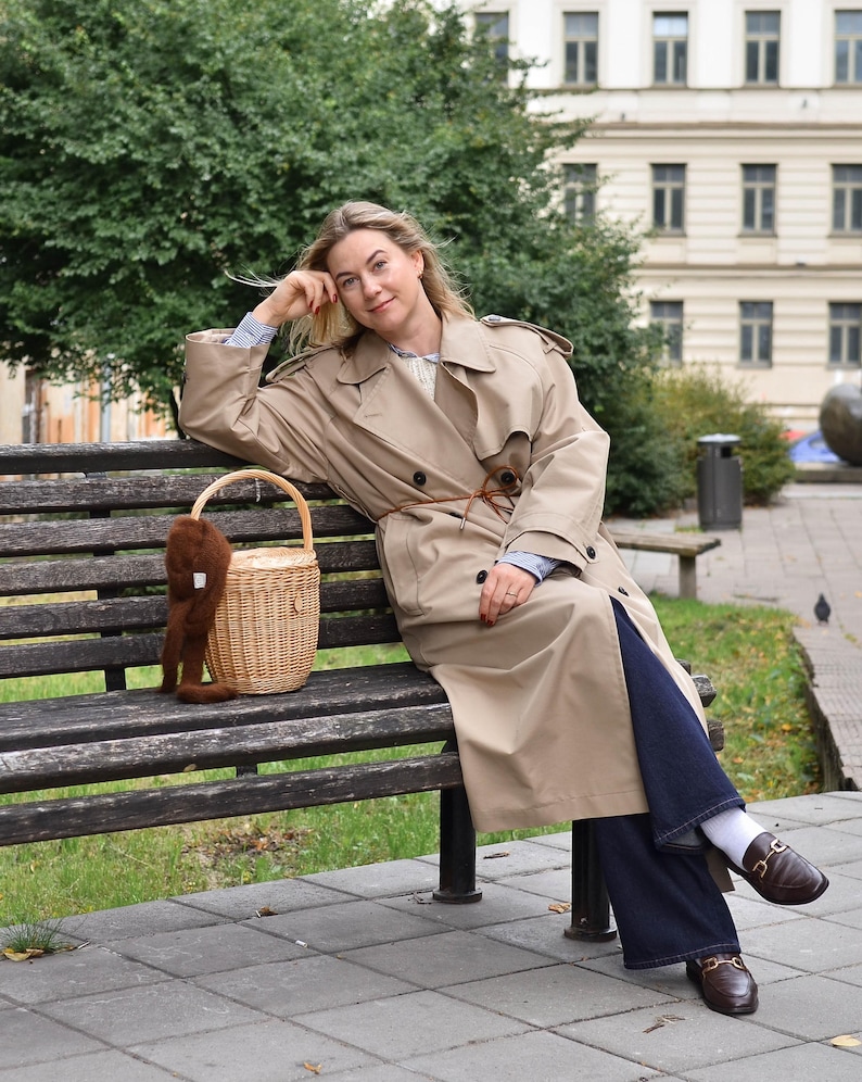 May include: A woman wearing a tan trench coat, blue jeans, and brown loafers sits on a wooden bench. A woven basket with a brown furry accessory rests beside her. The background includes a building and green trees.