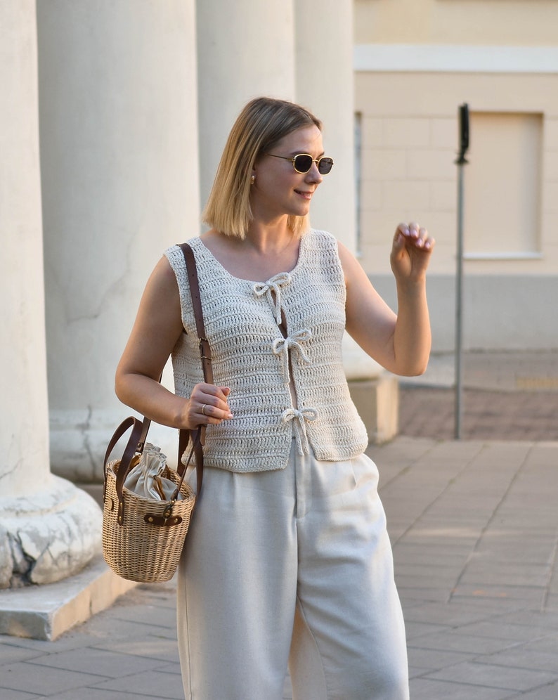 May include: A woman wearing a white crocheted sleeveless top with a tie at the front, white pants, and sunglasses. She is carrying a brown leather and wicker bag.