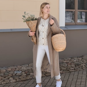 May include: A woman in a tan overcoat, white sweater, and white jeans, carrying a bouquet of flowers in brown paper and a wicker basket. She wears white trainers and gold jewellery. The background shows a building with a light wall and a stone path.