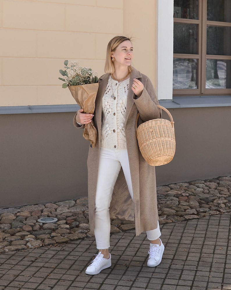 May include: A woman in a tan overcoat, white sweater, and white jeans, carrying a bouquet of flowers in brown paper and a wicker basket. She wears white trainers and gold jewellery. The background shows a building with a light wall and a stone path.