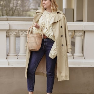 May include: A woman in a beige trench coat, cream-coloured knitted cardigan, and dark blue jeans, holding a woven basket. She wears black loafers. The background shows a white balustrade and a blurred outdoor scene.